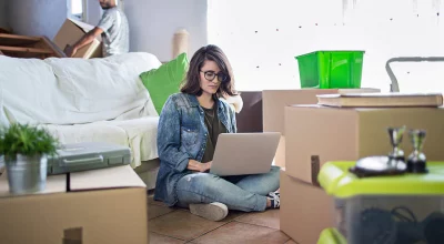 woman sitting on the floor working on a laptop surrounded by packing boxes.