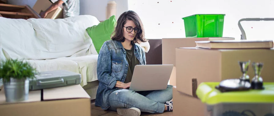 woman sitting on the floor working on a laptop surrounded by packing boxes.