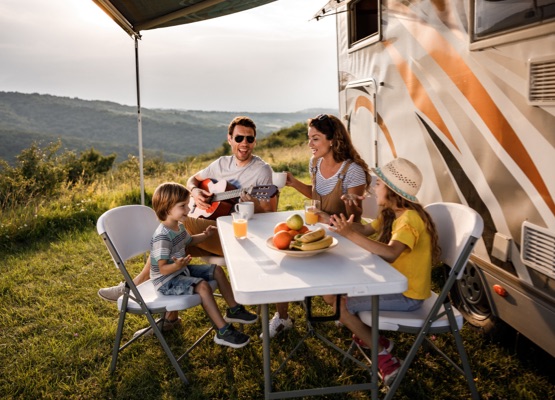 Happy family having fun while singing at picnic table by the camper trailer in nature. Man is playing a guitar.