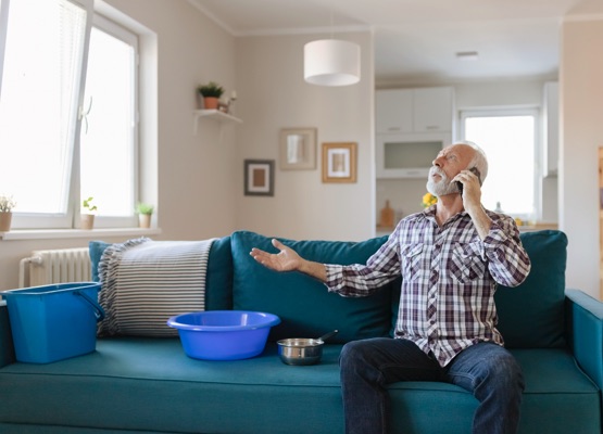 Angry Bearded Senior Man Talking on Smartphone Near Plastic Wash Bowl at Home in the Living Room Because of Roof Leaking