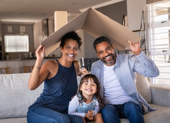 Portrait of smiling family sitting on couch holding cardboard roof and looking at camera. African and indian parents with funny daughter holding cardboard roof over heads while sitting on sofa in their new home. Happy mother and father with daughter in new house at moving day with copy space.