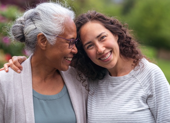 A senior woman embraces her millennial Eurasian daughter as they happily walk through a natural parkland area and enjoy their time together. The daughter is smiling at the camera.