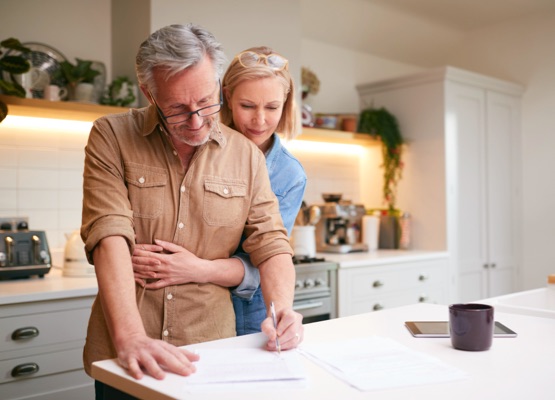 Mature Couple Reviewing And Signing Domestic Finances And Investment Paperwork In Kitchen At Home
