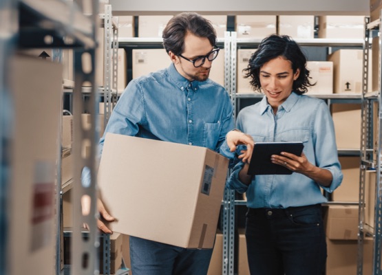 Female Inventory Manager Shows Digital Tablet Information to a Worker Holding Cardboard Box, They Talk and Do Work. In the Background Stock of Parcels with Products Ready for Shipment.