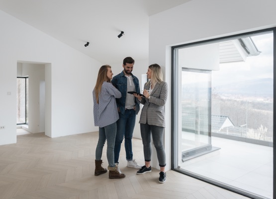 Real Estate Agent Shows Bright New Apartment to a Young Couple. Successful Young Couple Becoming Homeowners, Embraces and Hugs Each Other. Spacious Bright Home with Big Windows