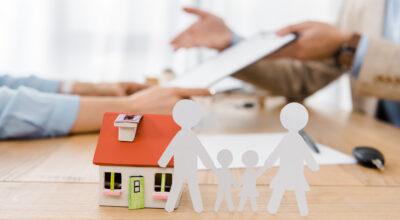 White paper cut family and house model on wooden table with blurred people at background, life and house insurance