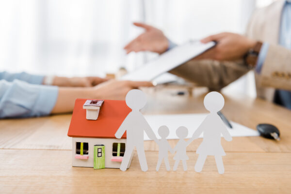 White paper cut family and house model on wooden table with blurred people at background, life and house insurance