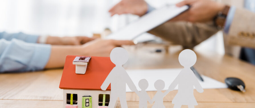 White paper cut family and house model on wooden table with blurred people at background, life and house insurance